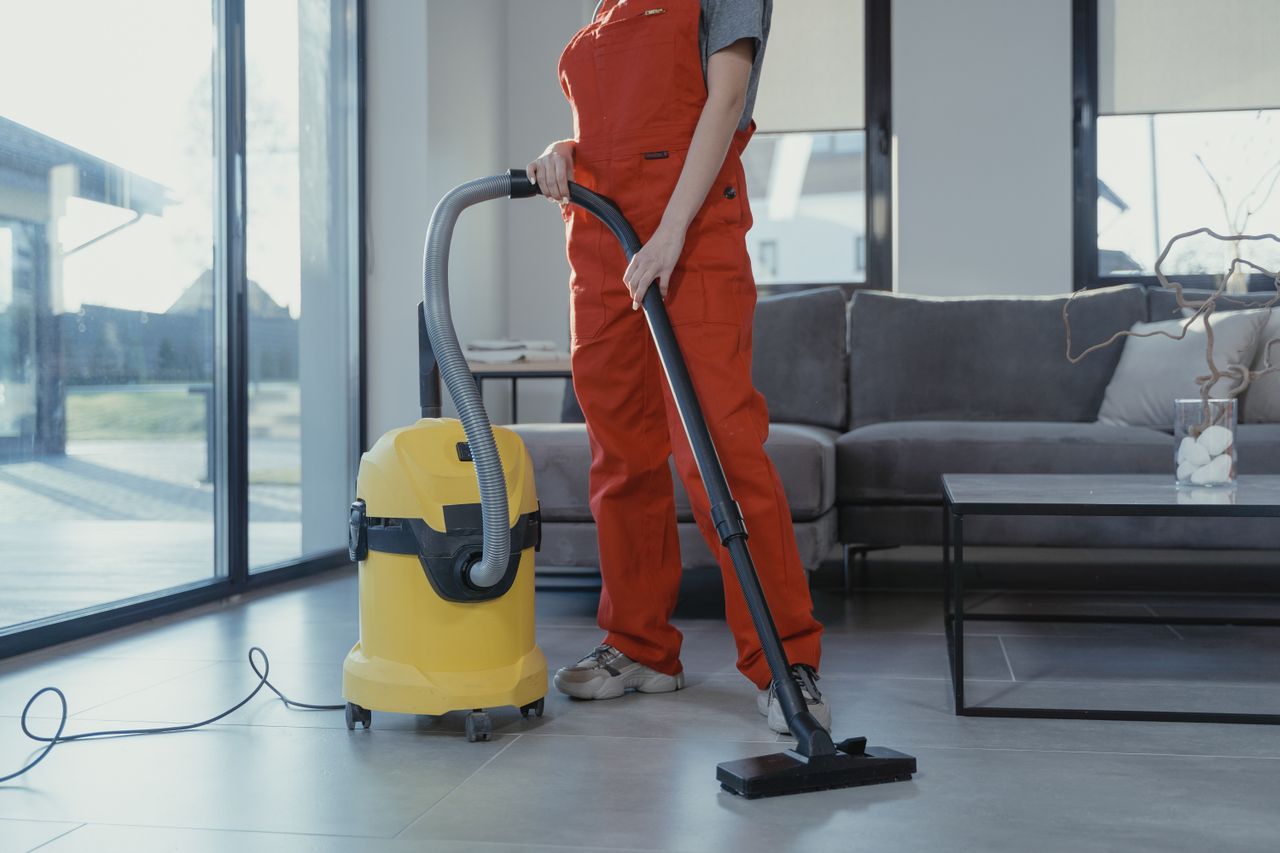 services-03 A professional cleaner in red coveralls vacuuming a modern living room with a yellow vacuum cleaner.